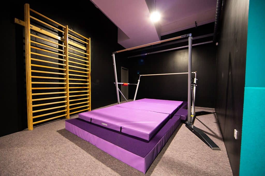 Gymnastics training area with purple tumbling mats, a horizontal bar, and wooden wall bars for strength and flexibility exercises. Suitable for gymnastics practice and physical therapy.