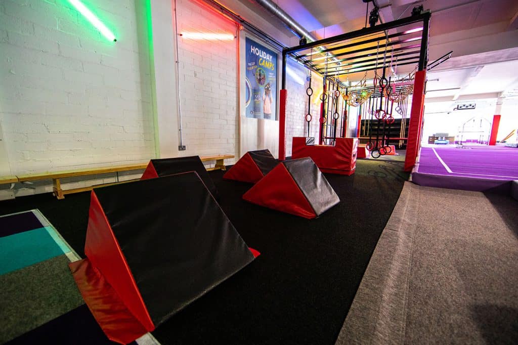 Bright indoor fitness area with padded climbing equipment, gymnastic rings, and sports mats for kids' recreational activities. Vibrant lighting and colourful decor create an energetic atmosphere.