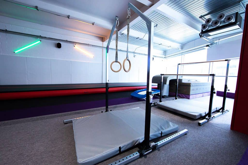 Gymnastics training area with rings, mats, and safety equipment in a modern fitness facility with colourful LED lighting and high ceilings.