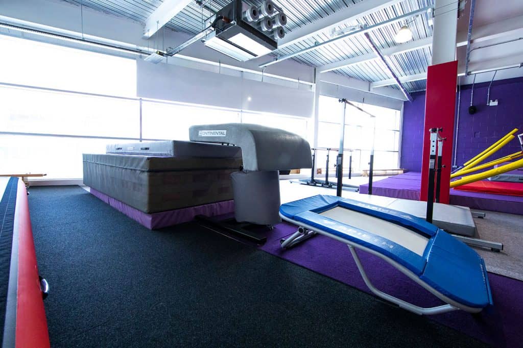A spacious indoor gymnastics training facility with various equipment including a trampoline, foam pit, and padded mats, illuminated by large windows and featuring vibrant purple and red accents.