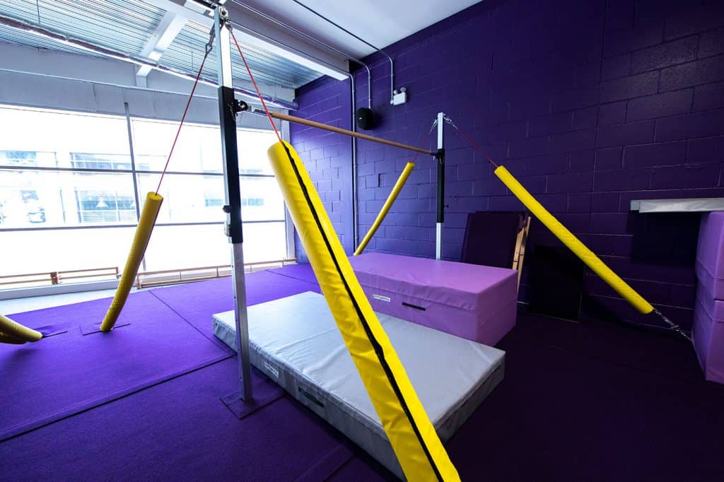 Padded gymnastics area with purple mats, yellow foam safety bars, and a vaulting box in a brightly lit indoor sports facility with large windows and purple brick walls.