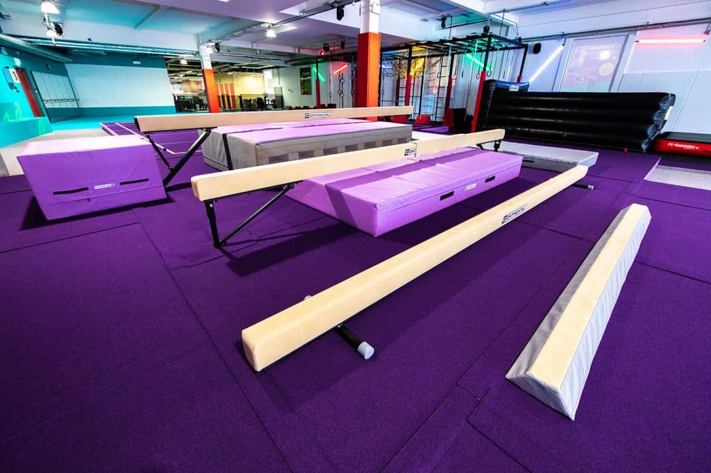 Portable gym equipment, gymnastics beams, and yoga mats on purple floor in modern indoor fitness centre. Well-lit space for various workout activities and training sessions.