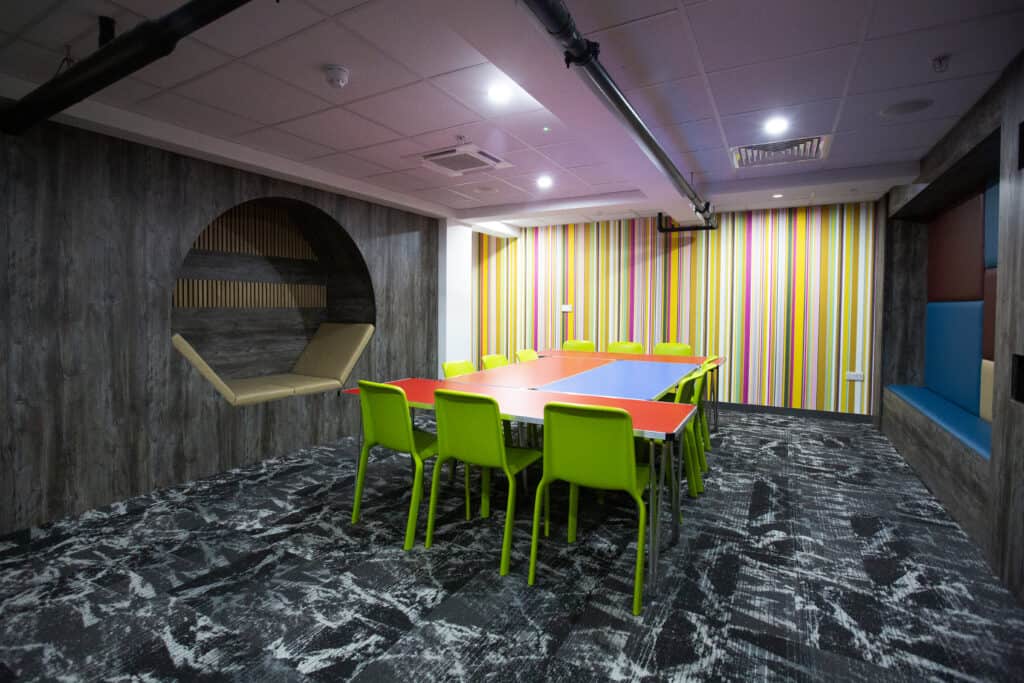 Brightly coloured meeting room with a large rectangular table surrounded by lime green chairs, vibrant striped wallpaper at the back, a patterned carpet, and cozy seating nook on the wall.