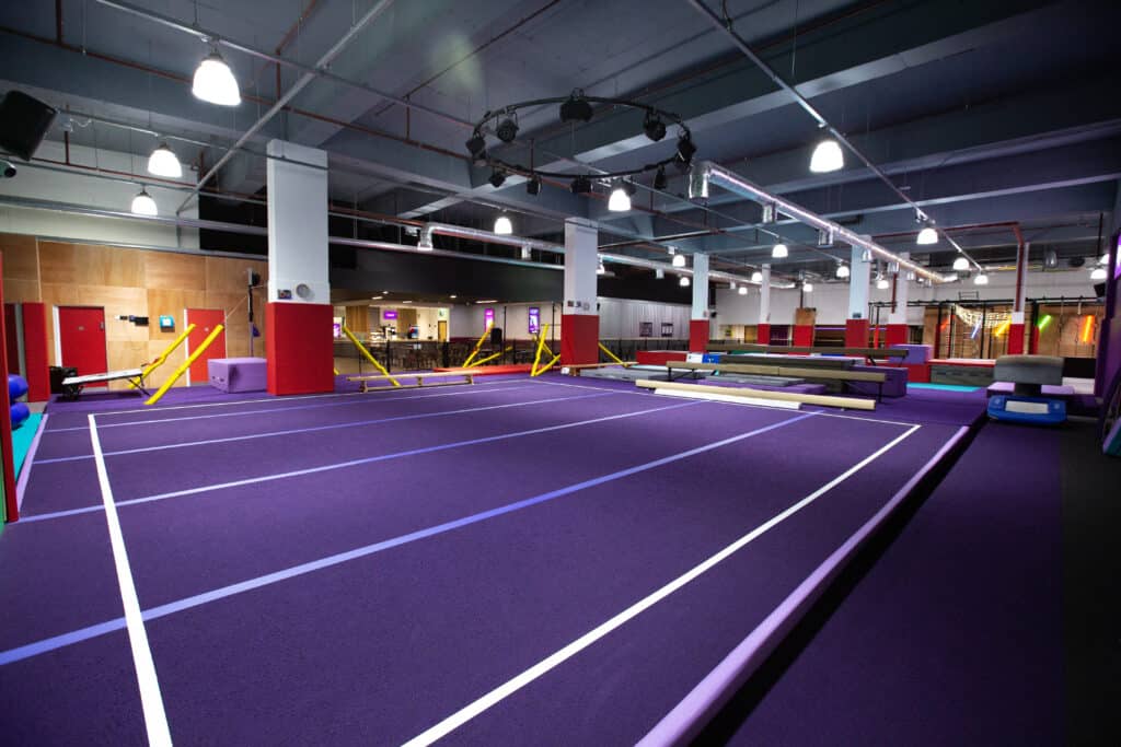 Purple indoor trampoline park with various soft play equipment and safety padding, illuminated by bright ceiling lights, in a modern entertainment centre setting.