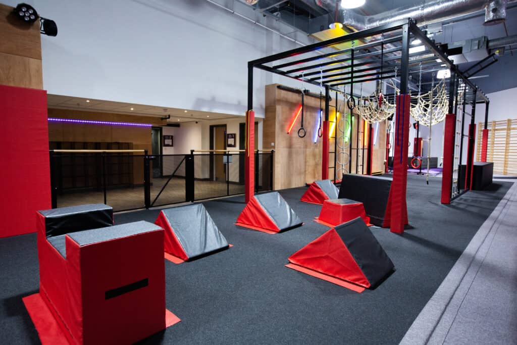 Bright indoor adventure and obstacle course equipment in a modern gym or fitness centre, featuring padded climbing blocks, rings, ropes, and neon lighting for functional training and fitness activities.