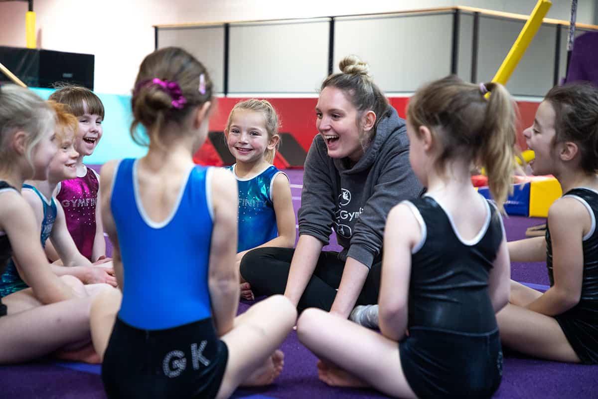 Energetic young gymnasts sitting in a circle during a training session with their smiling coach in a colourful indoor gymnastics facility.