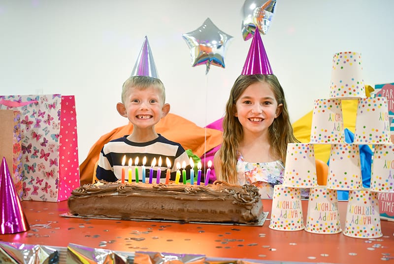 Children celebrating birthday with chocolate cake, party hats, balloons, and colourful decorations at a festive party.