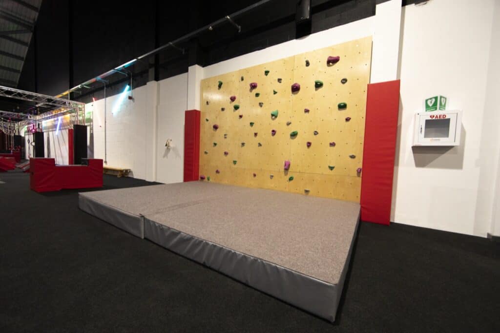 Climbing wall indoors with padded floor and emergency AED on the wall for safety.