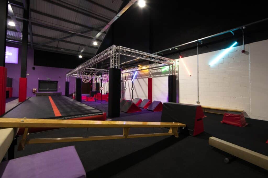 Climbing gym with an indoor obstacle course featuring a raised ramp, hanging rings, and various soft play elements under colourful LED lights for enhanced training and recreation.