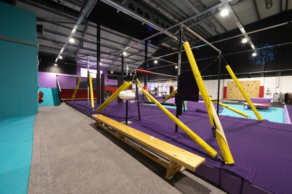 Climbing gym indoor training area with purple padded flooring, yellow padded beams, and various climbing structures for children and beginners. Bright, spacious, and well-lit with high ceilings.