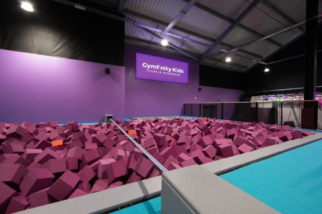 Pink foam pit filled with foam blocks at Gymfinity Kids indoor trampoline park, purple wall with Gymfinity Kids branding, high ceiling with industrial lighting.