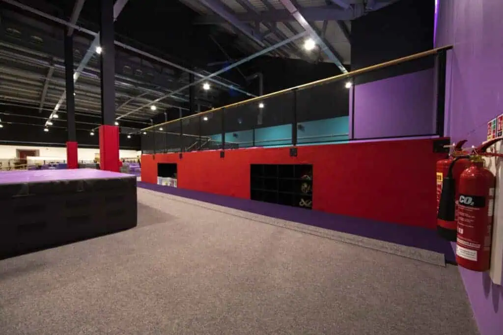 Foam pit area with padded flooring, red padded walls, and black storage compartments in an indoor trampoline or ninja warrior park. Safety equipment like fire extinguishers are visible on the wall.