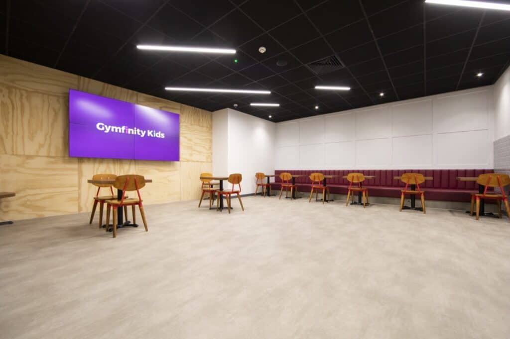 Gymfinity Kids indoor play area with modern decor, wall-mounted screen, wooden accents, and ample seating, creating a welcoming environment for children to enjoy active fun and activities.