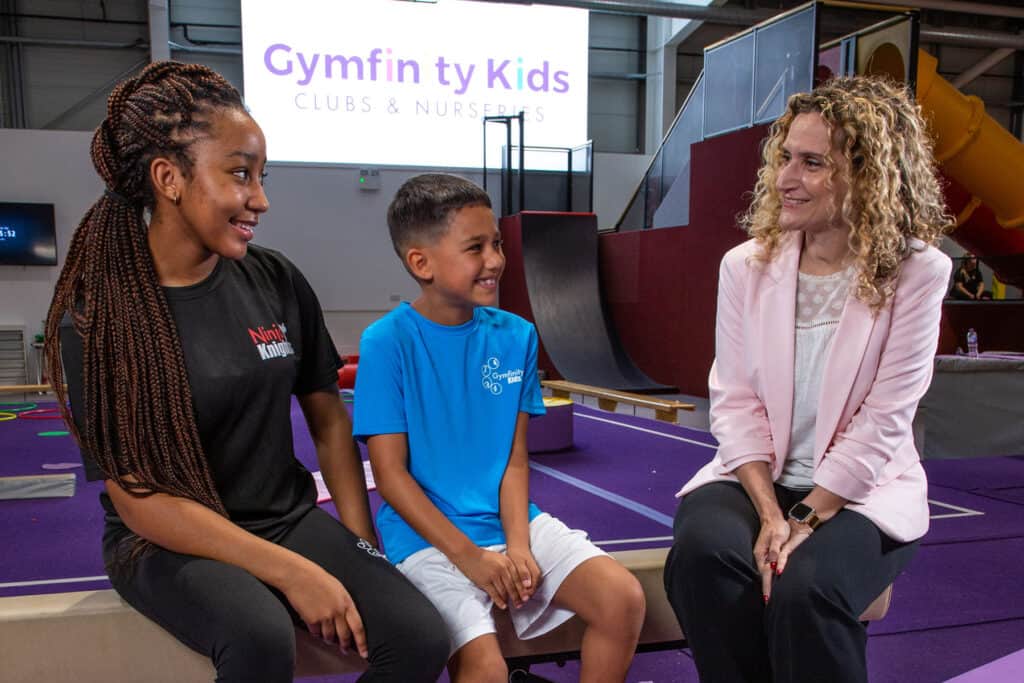 Children and a woman sitting and talking in a gymnastics training centre with gymnastics equipment in the background.