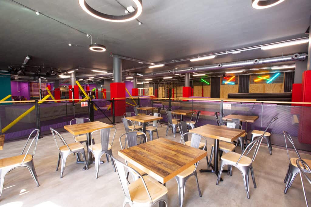 Modern indoor trampoline park with seating area, wooden tables, metal chairs, colourful padded zones, neon lights, and safety padding for a fun and active experience.