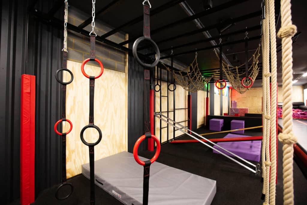 Climbing and gymnastics equipment in a modern indoor adventure play area with ropes, rings, and padded mats for active children's play and exercise.