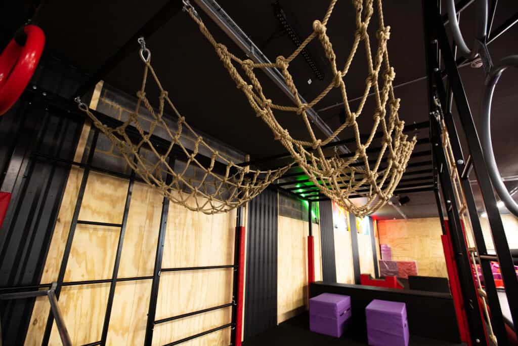 Climbing net obstacle for children in an indoor play area with colourful walls and padded seating.