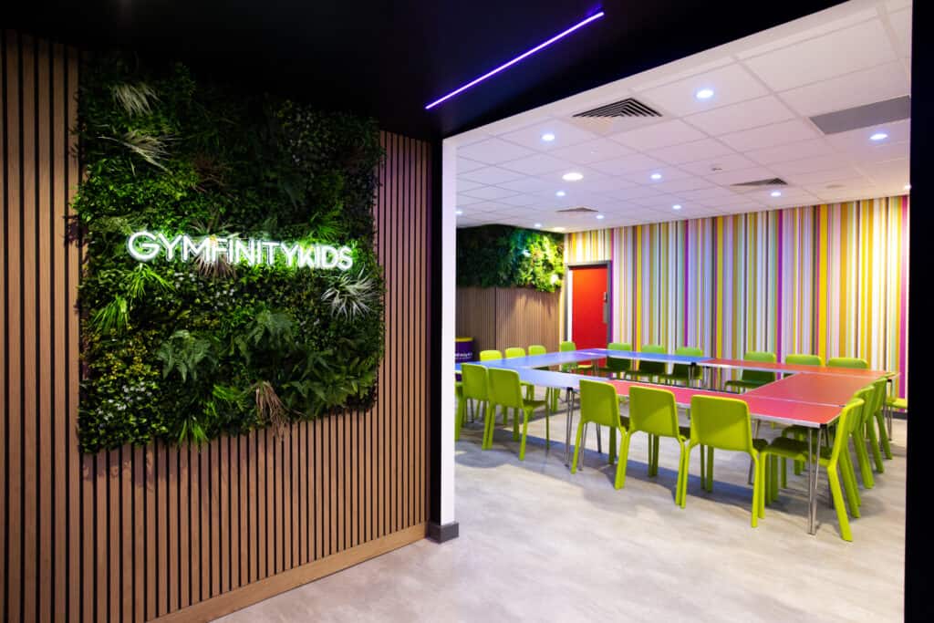 Brightly coloured kid’s activity room with green chairs, pink tables, striped wallpaper, and a lush green wall with "GYMFINITYKIDS" signage illuminated in neon.