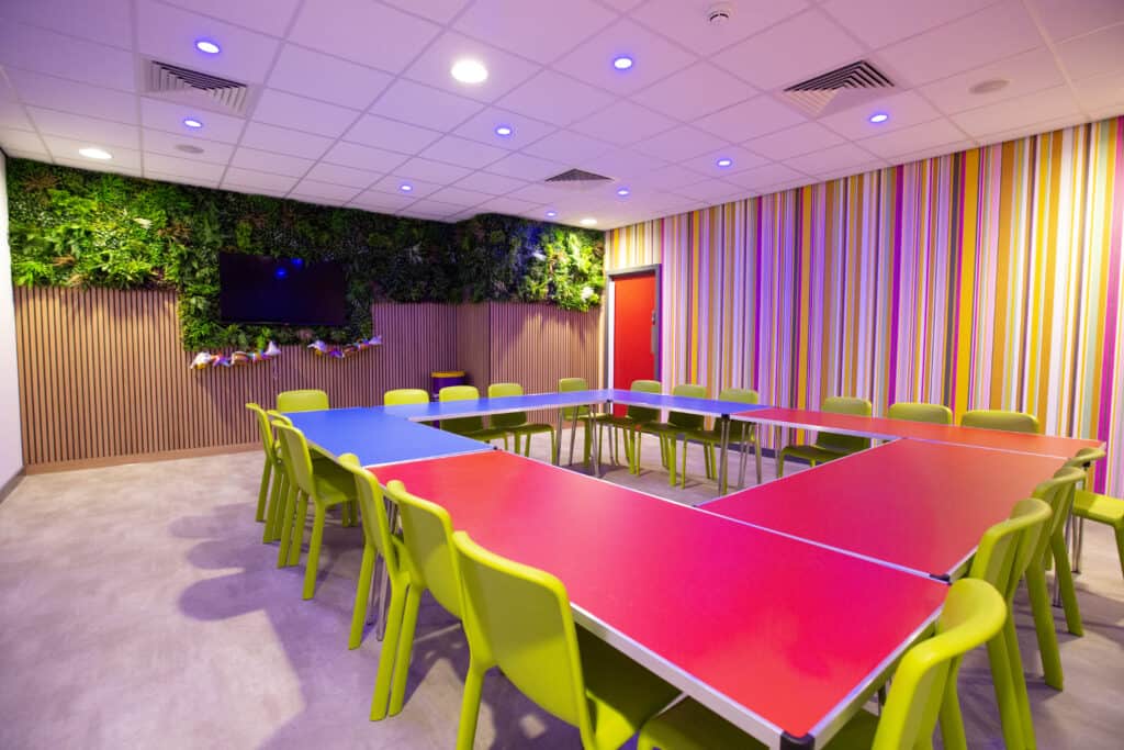 Brightly coloured meeting room with red, blue, and pink tables, lime green chairs, a green wall with plants, and colourful striped wallpaper, perfect for creative brainstorming sessions.