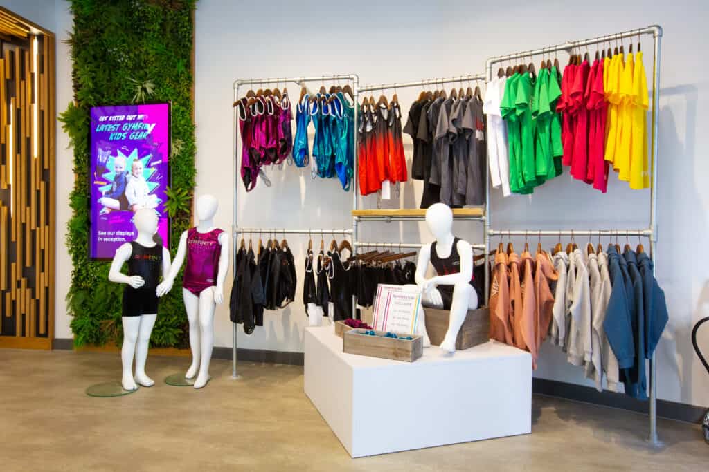 Brightly coloured children's gymwear displayed on racks and mannequins inside a sporting goods store. The setup features various t-shirts, shorts, and tank tops in vibrant colours, with promotional digital signage nearby.