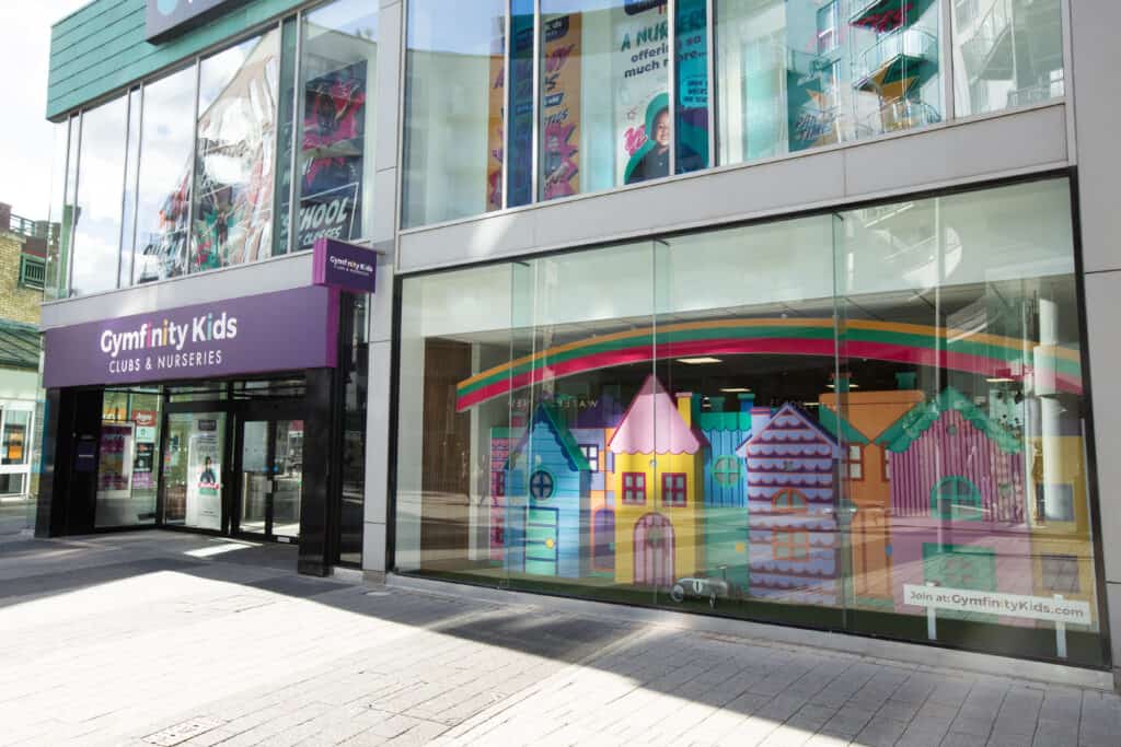 Colourful children's playhouse models inside Gymfinity Kids nursery through large glass windows on a modern city building.
