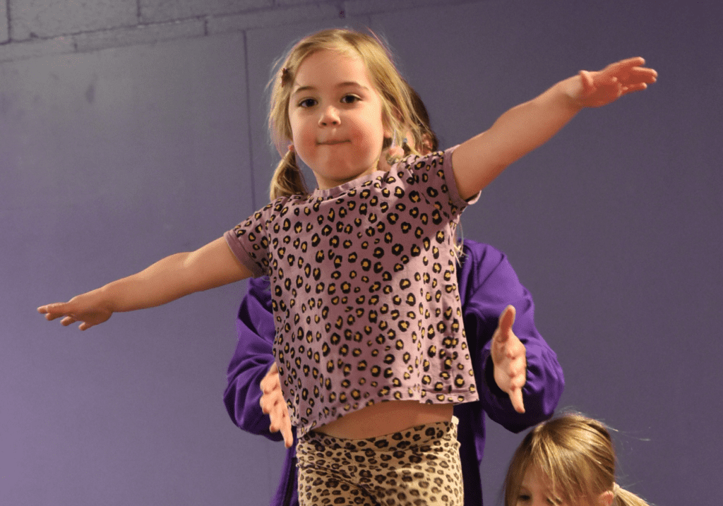 Child girl with blonde hair in pigtails wearing animal print outfit, balancing with arms outstretched on stage.