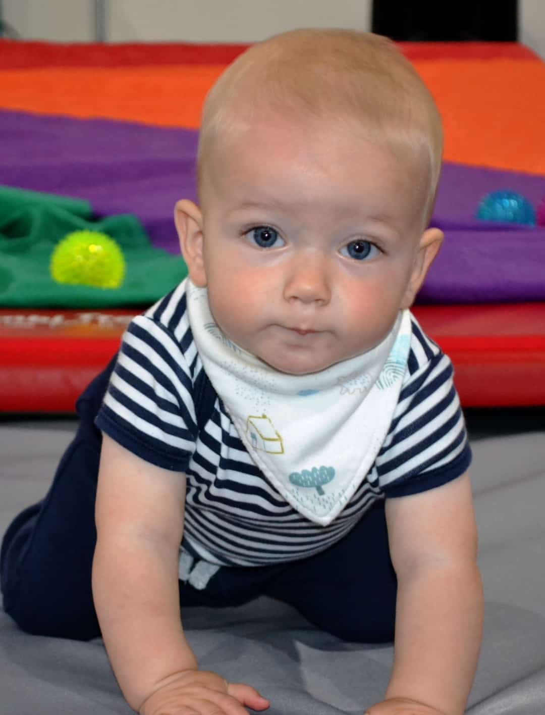 Baby boy crawling on the floor with a colourful play mat and toys in the background, wearing a striped t-shirt and a bib, showcasing early childhood development and playtime.