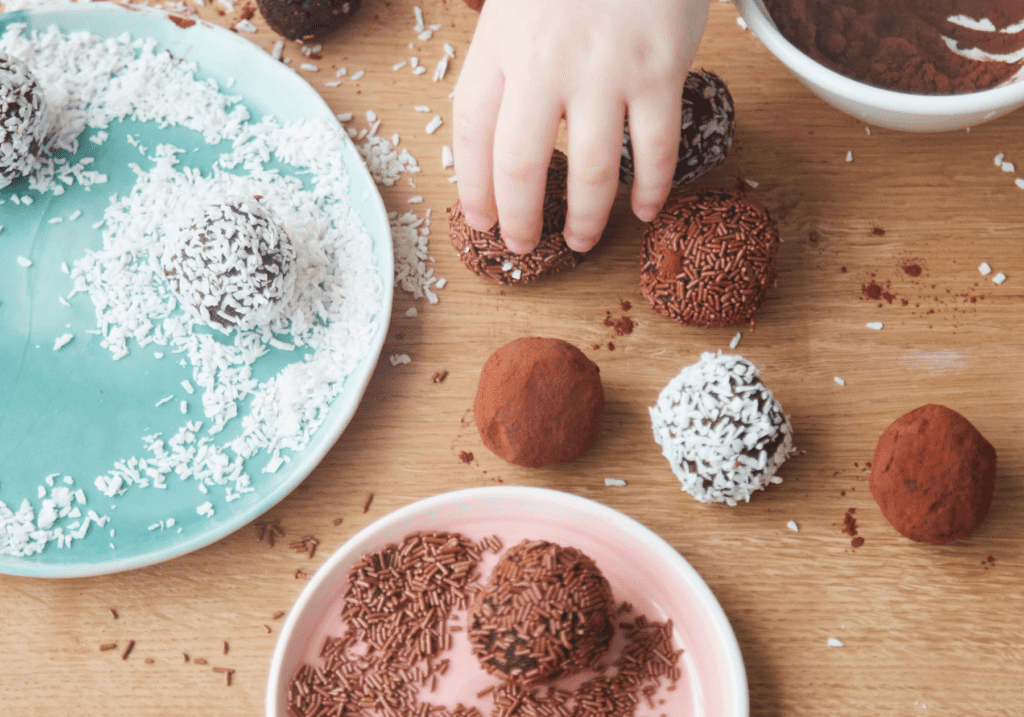 Rich chocolate truffles being coated in chocolate sprinkles and shredded coconut on a wooden surface. Hand is seen placing a truffle into a bowl of sprinkles for coating. Various bowls of toppings and finished truffles surround the workspace.