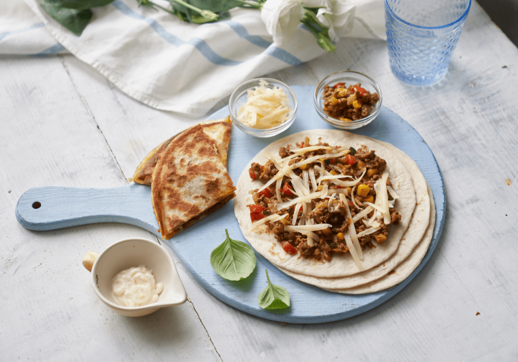 Savoury beef and vegetable filling on a soft wrap with shredded cheese, served with a side of naan bread, sour cream, and fresh basil leaves on a rustic blue wooden board.