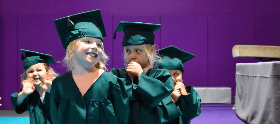 Graduating children in green caps and gowns during a school ceremony on a stage with a purple background.