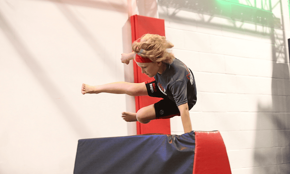 High-impact gymnastics training with a young girl performing a vault move over padded mats in an indoor gymnasium.