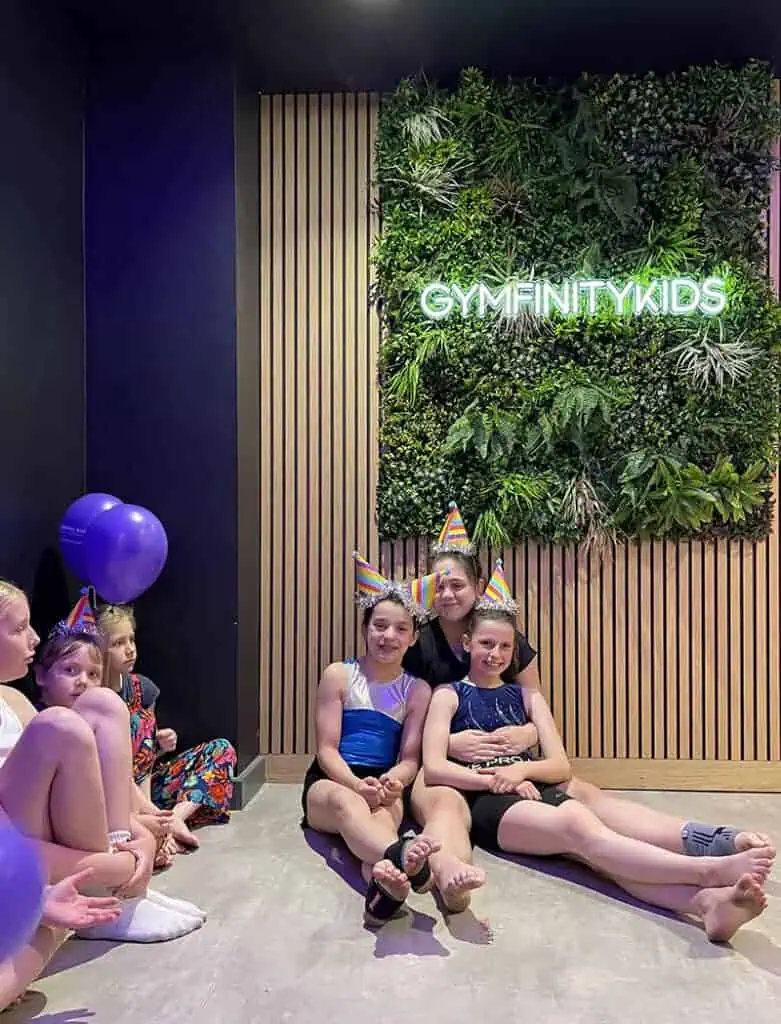 Gymfinitykids children’s gymnastics class celebrating with birthday hats at a vibrant indoor facility featuring a lush green wall and playful atmosphere.