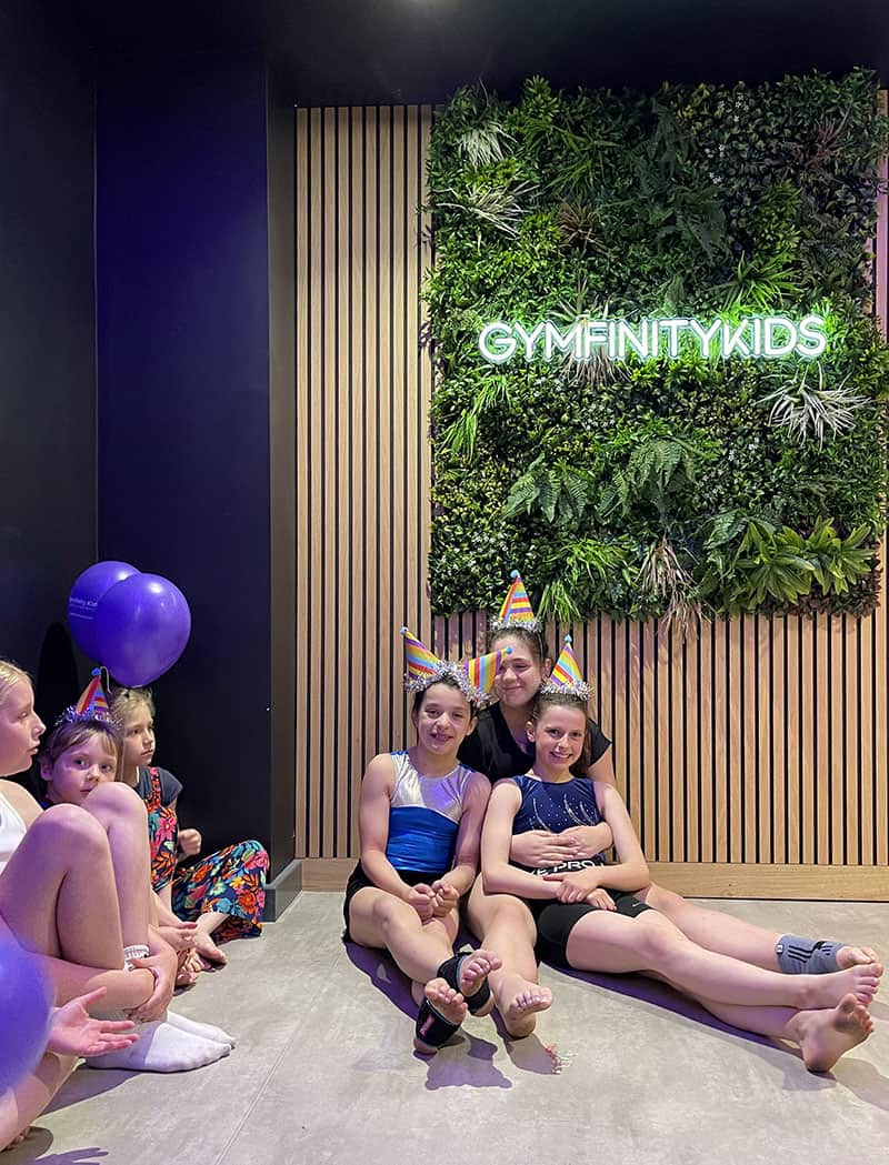Gymfinitykids children’s gymnastics class celebrating with birthday hats at a vibrant indoor facility featuring a lush green wall and playful atmosphere.