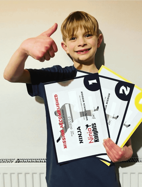Boy holding ninja certificates and giving a thumbs up, celebrating success in a fun activity or competition.