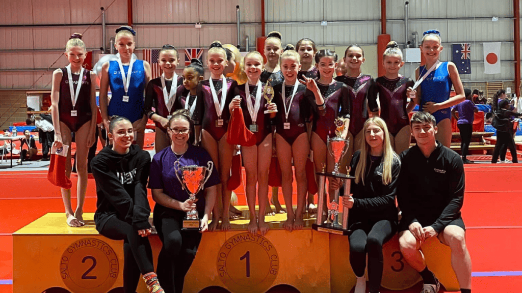 Girls' artistic gymnastics team celebrating victory at a competition, standing on winners' podium with gold, silver, and bronze medals, holding trophies, in an indoor gymnasium.