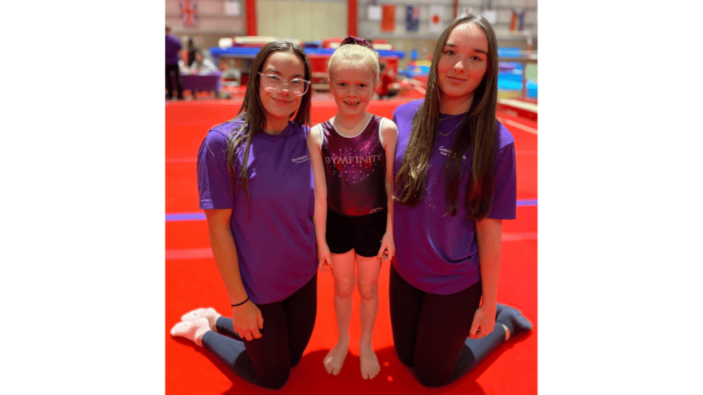 Gymnastics training session with two coaches and a young gymnast on a red mat, holding a trophy award in a colourful indoor sports facility.