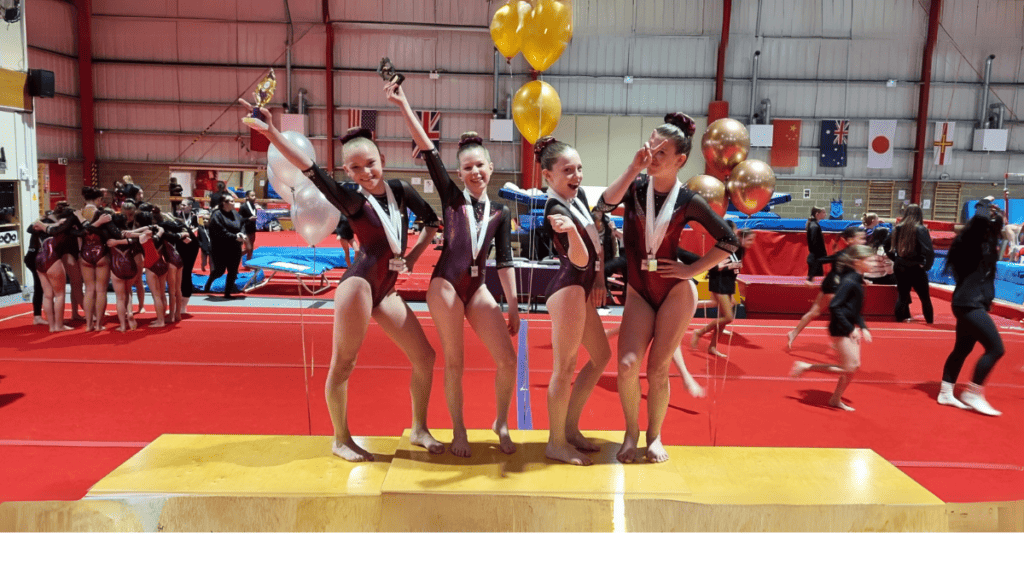 Gymnastics training session with young girl performing on balance beam in indoor gym, surrounded by coaches and banners, British flags visible in background.