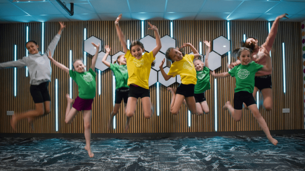 Children doing gymnastic exercise indoors, kids in colorful sportswear participating in a fitness activity, fun, energetic, and inclusive environment in a modern gym or sports centre, smiling and engaging in physical activity, learning teamwork and fitness.