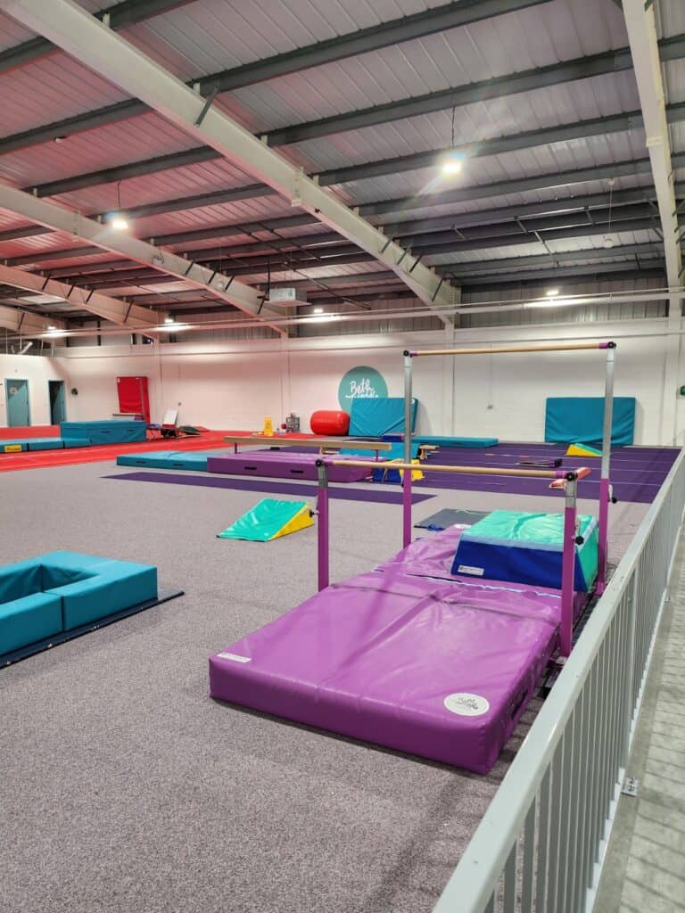 Bright indoor gymnastics training facility with colourful mats, bars, and equipment for children and athletes, featuring spacious area and high ceilings.