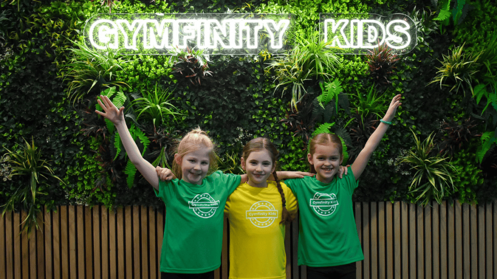 Gymfinity kids, children in matching green and yellow t-shirts smiling with arms raised, indoor gym facility with lush green plant wall featuring neon sign.
