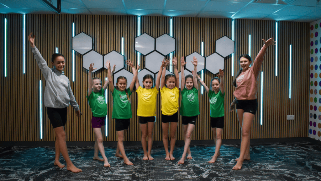 Children doing gymnastic exercise indoors, kids in colorful sportswear participating in a fitness activity, fun, energetic, and inclusive environment in a modern gym or sports centre, smiling and engaging in physical activity, learning teamwork and fitness.