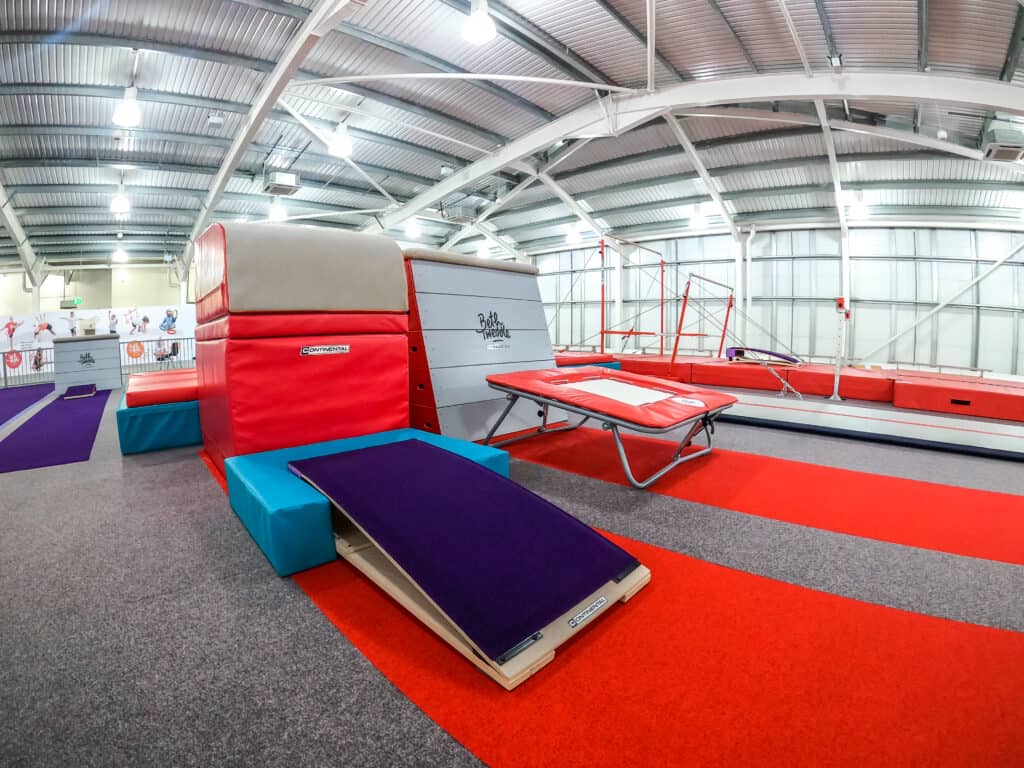 High-performance indoor gymnastics training arena with a springboard, foam pits, and balance beam, featuring vibrant red, purple, and grey safety padding in a spacious sports facility.
