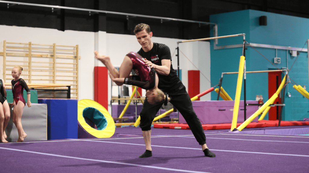 Young gymnasts practising at an indoor gymnastics training facility with coaching help, colourful training equipment, and purple mats.