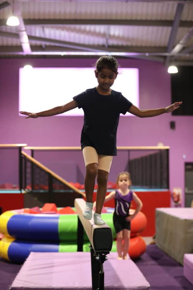 Children practicing balance on a slackline inside an indoor play centre with soft play equipment and colourful surroundings.