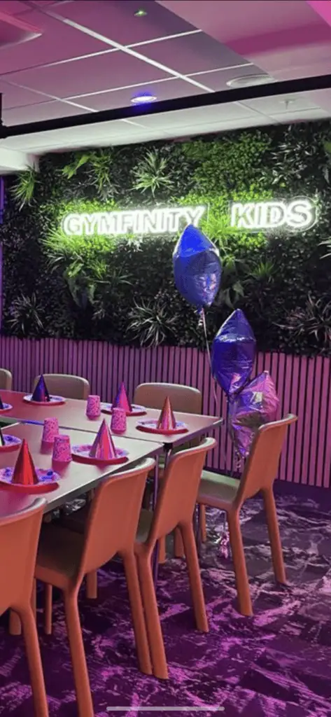 NEON sign with "GYMFITTY KIDS" illuminated in white on a green leafy wall, decorated with purple and blue balloons, pink party hats, and candles, set up for a children's birthday celebration.