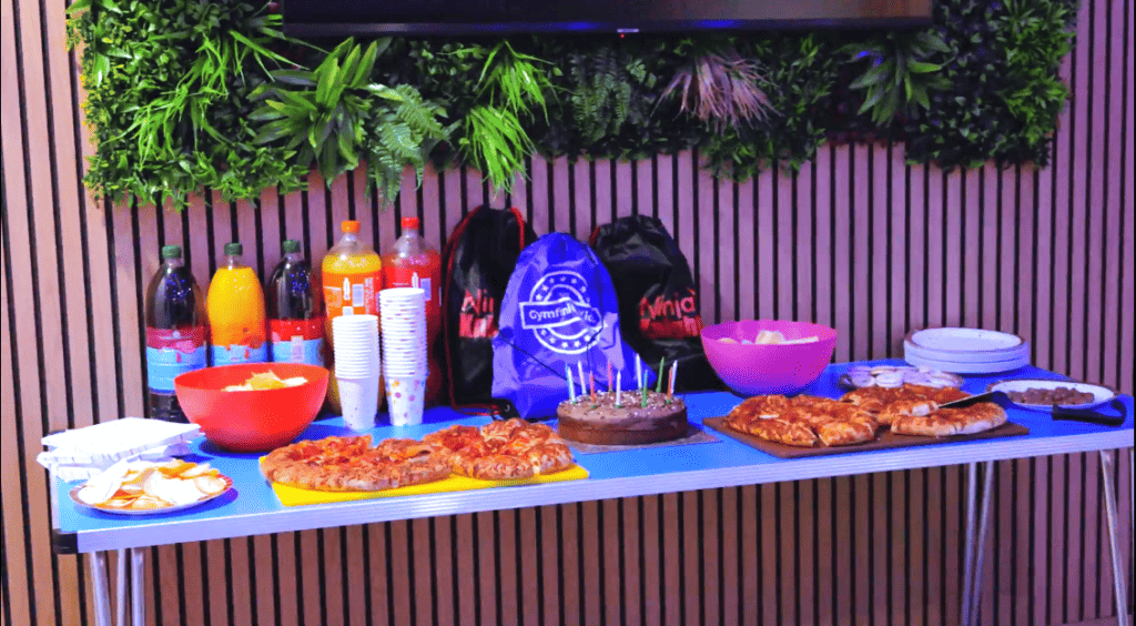 Pizza, snacks, and beverages set up on a table for a celebration with green foliage decor behind.