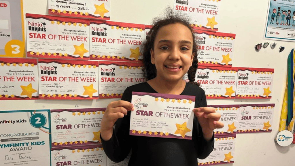 Star of the week award certificate held by a smiling young girl in front of a wall decorated with multiple "Ninja Knights" achievement certificates, celebrating her accomplishments at school.