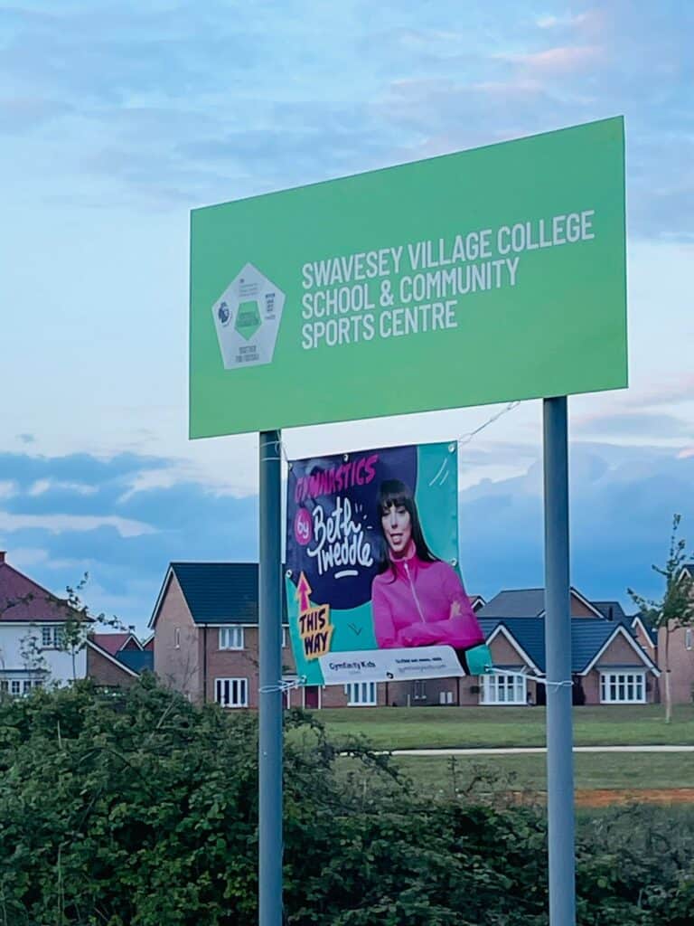 Bright green signboard for Swavesey Village College School & Community Sports Centre with a promotional poster for gymnastics by Betfo Tweddle.