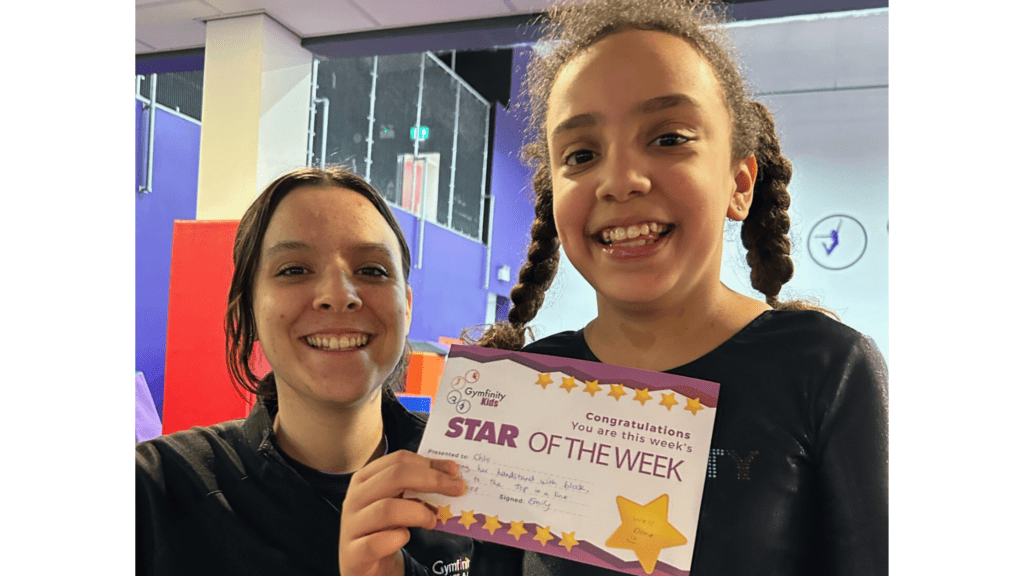 Brightly smiling young girl holding a "Star of the Week" certificate with an adult woman at a gymnastics centre, celebrating achievement and success.