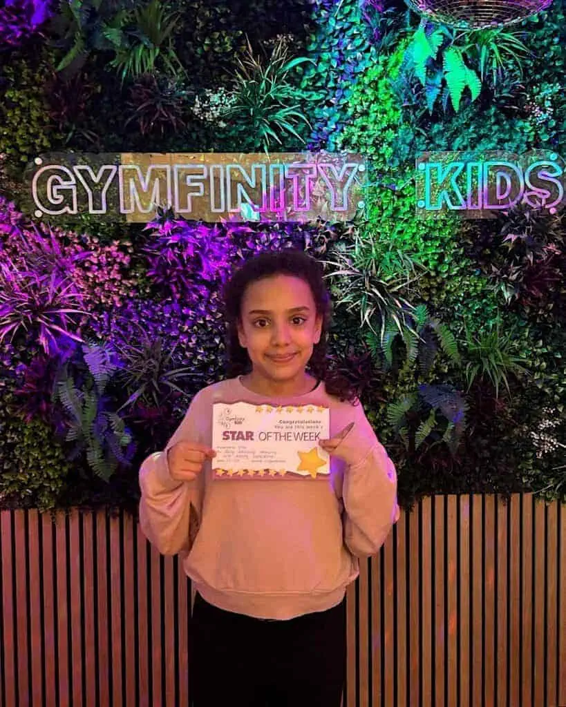 Young girl holding an award certificate in front of a vibrant green wall with neon signs reading "GYMFINITTY KIDS".